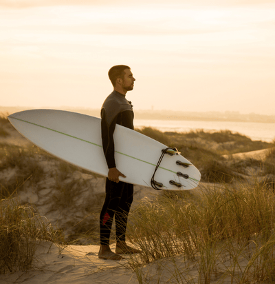 A man in the dunes with a swimsuit and a surf board under his arm watching the waves at sunset
